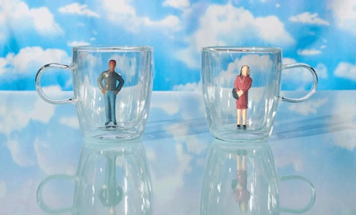 Two small figurines, one masculine and one feminine, perched within glass mugs, with a backdrop of clouds on a blue sky
