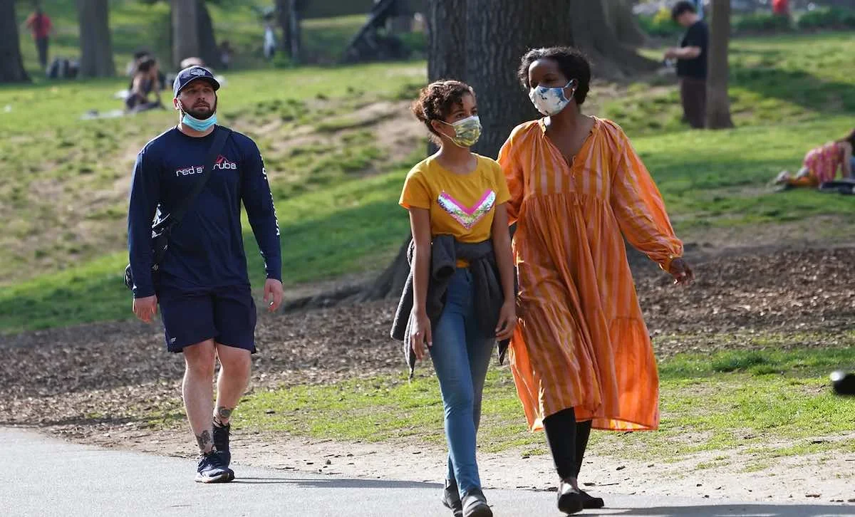 Photograph of people walking in a park, some with masks, one with a mask pulled down to his chin