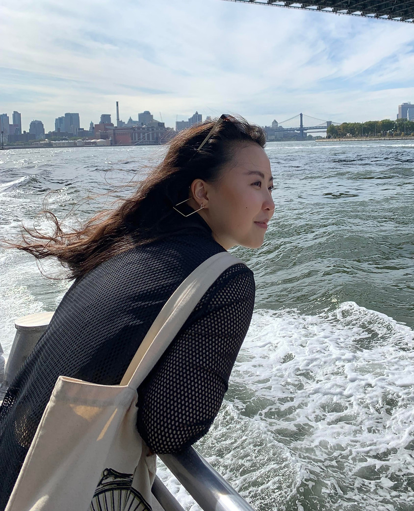 A Korean-Canadian person looks off the the right, leaning over a boat's railing, a canvas tote slung over their shoulder. In the distance, the New York City skyline is visible.